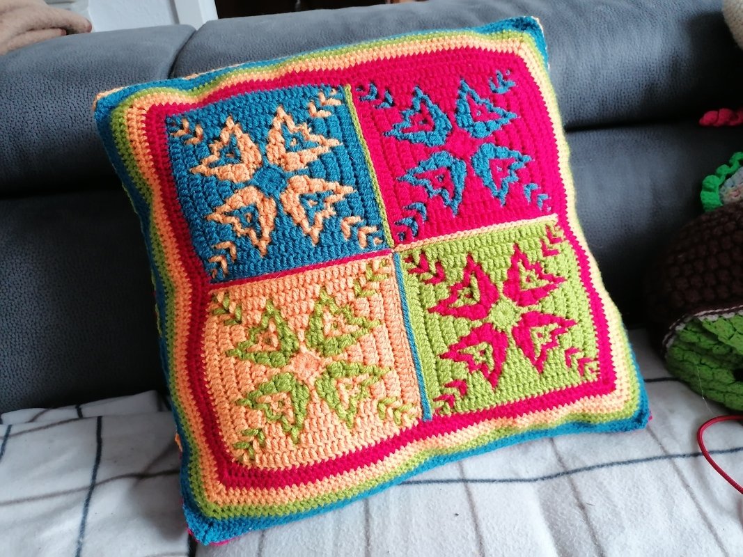 Gehäkeltes Kissen mit vier bunten Quadraten in Blau, Pink, Orange und Grün auf grauem Sofa