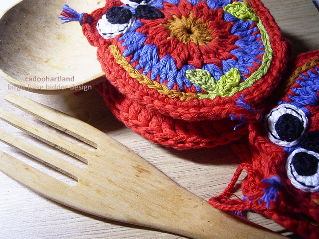 Nahaufnahme von zwei gehäkelten Topflappen in Rot mit blauem, grünem und gelbem Muster und weißen sowie schwarzen Augen, neben einem Holzlöffel und einer Holzgabel.