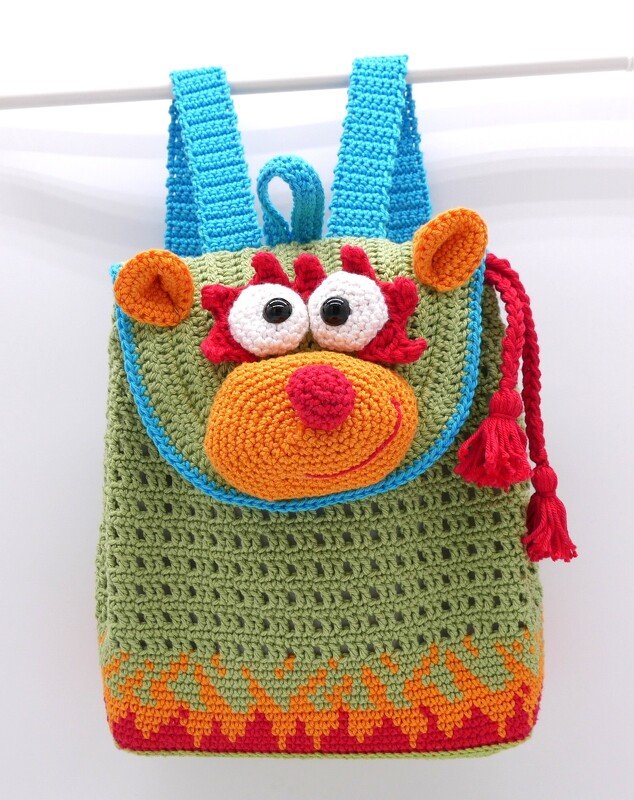 Gehäkelter Rucksack in Grün mit blauen Trägern, einem Tiergesicht mit großen Augen, orangefarbener Schnauze und roter Quaste