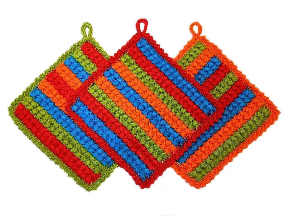 Drei bunte gestreifte Topflappen mit Aufhängeschlaufen, gehäkelt in Rot, Orange, Blau und Grün auf weißem Hintergrund.