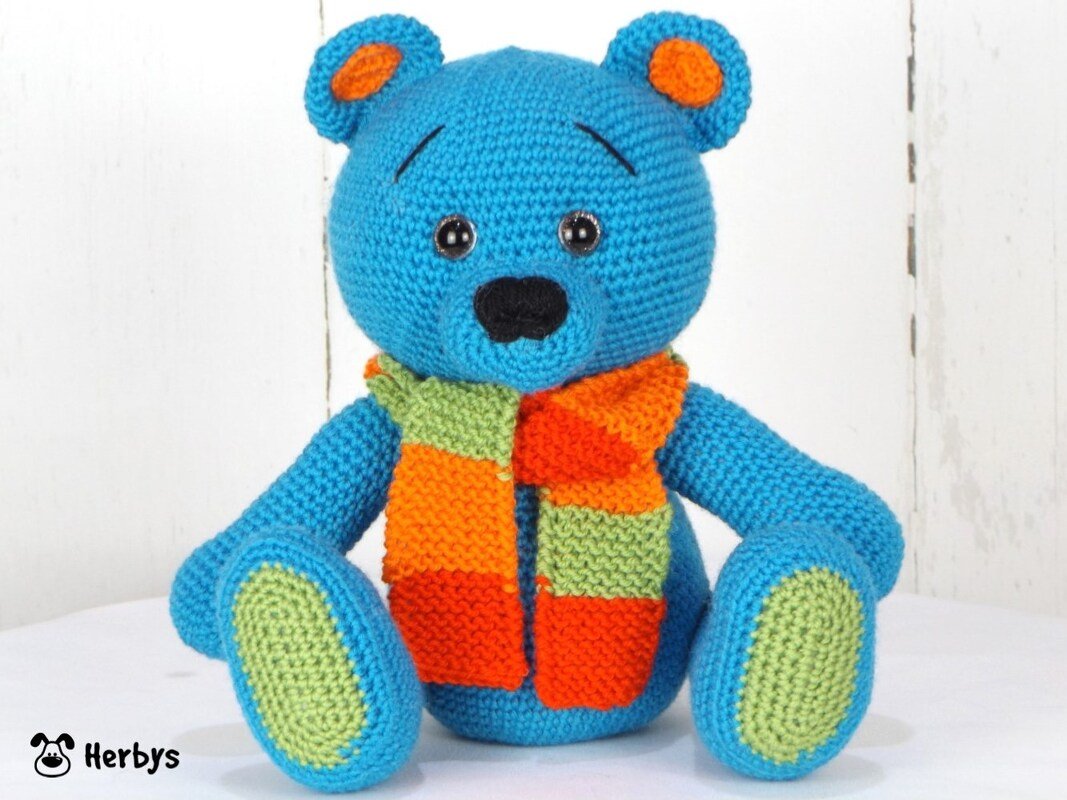 Bright blue crocheted teddy bear with orange and green striped scarf and green paw pads sitting on a white surface