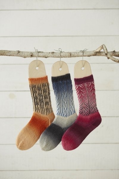 Drei handgestrickte Socken mit Farbverlauf und unterschiedlichen Zopf- und Strukturmustern an einem Ast.