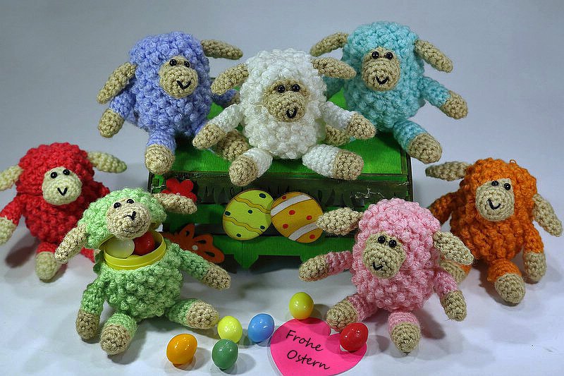 Sieben bunte, gehäkelte Amigurumi-Schäfchen sitzen um Ostereier und bunte Schokolinsen; ein rosa Herz mit „Frohe Ostern“ liegt davor.