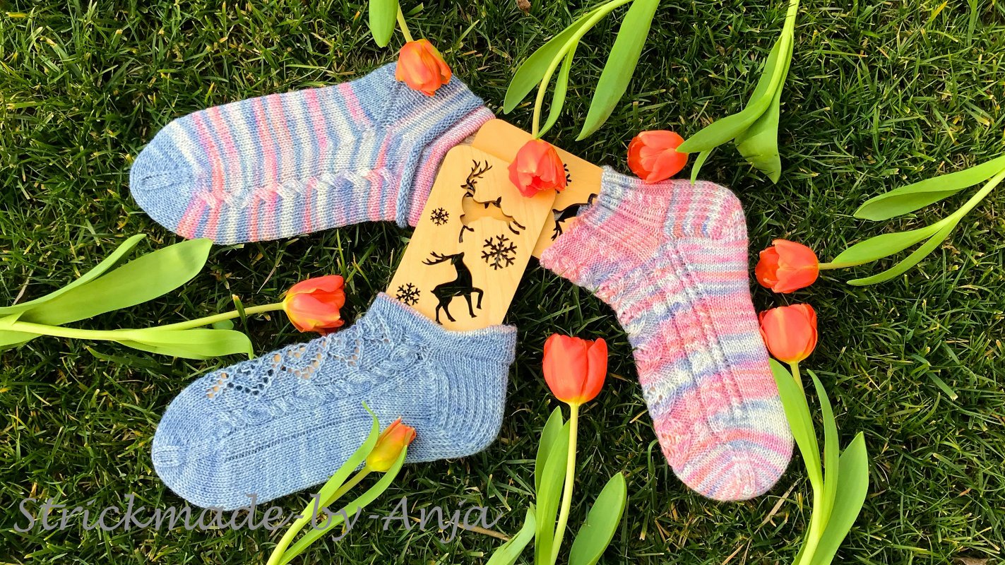 Drei kurze gestrickte Socken mit Loch- und Streifenmustern auf Holzsockenblockern, umgeben von roten Tulpen auf Gras