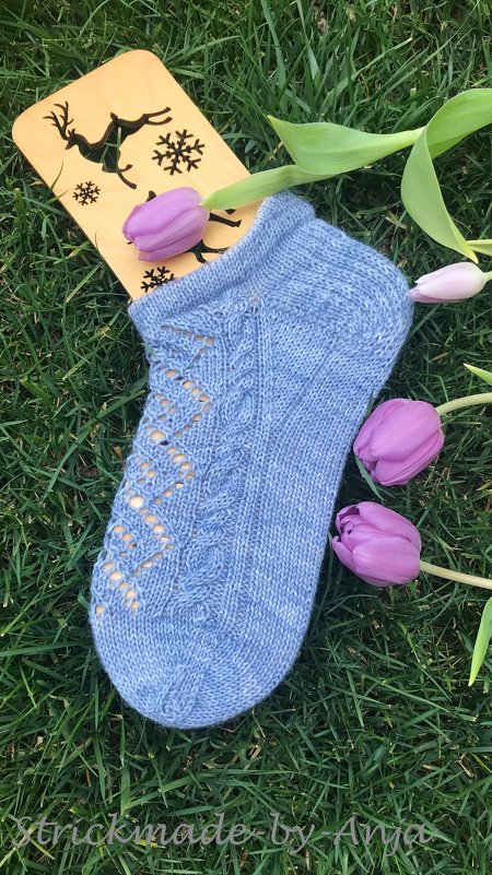 Hellblaue gestrickte Kurzsocke mit Loch- und Zopfmuster auf Gras neben lila Tulpen und Sockenbrett