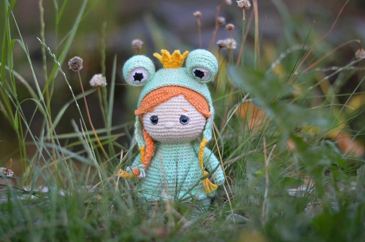 Gehäkelte Amigurumi-Puppe mit mintgrüner Froschkapuze, orangen Zöpfen und kleiner Krone sitzt im Gras