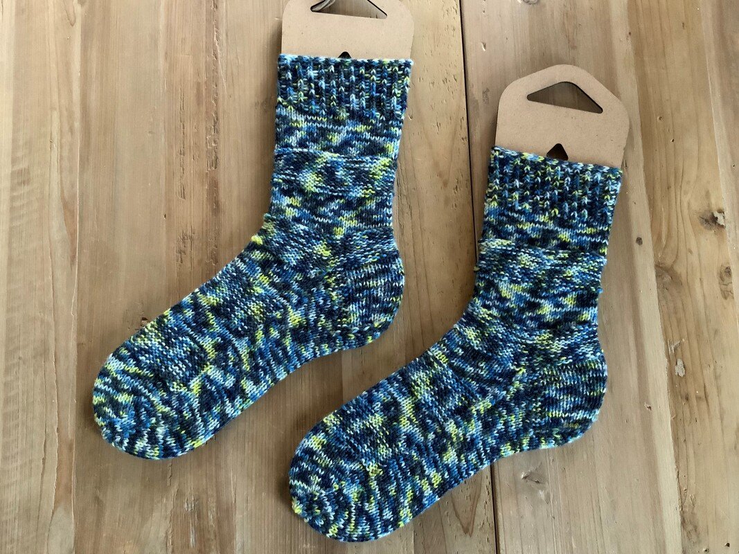 Zwei blau-gelb gemusterte Stricksocken auf hellem Holz mit braunen Etiketten.