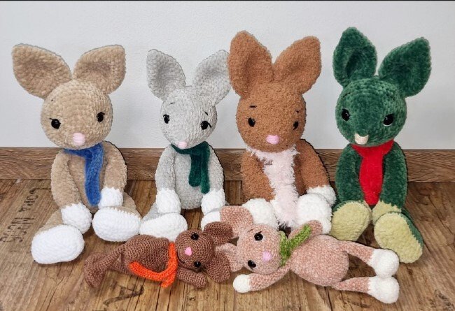 Vier sitzende gehäkelte Amigurumi-Hasen mit Schals und zwei kleinere Hasen davor auf Holzboden
