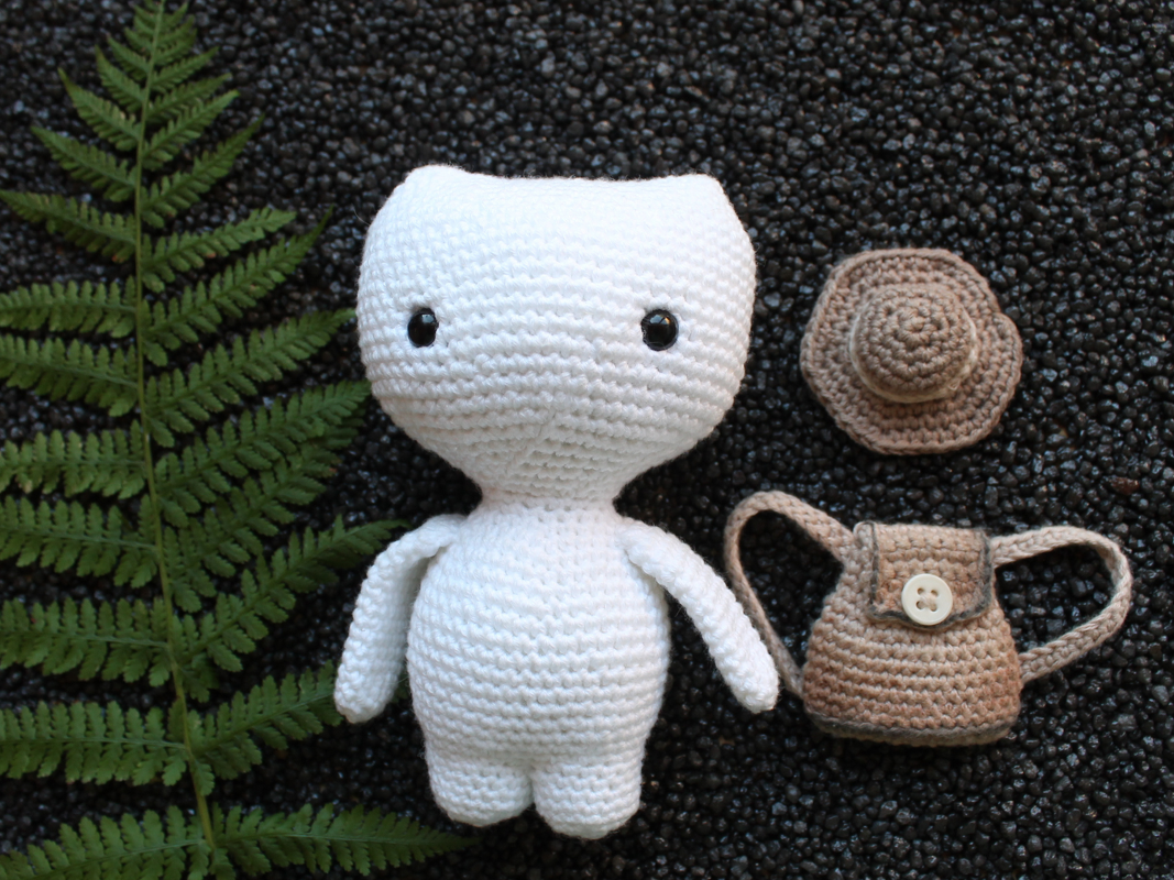 Weißes gehäkeltes Amigurumi-Kätzchen mit schwarzen Augen, brauner Mini-Hut und kleiner Tasche, Farnblatt links