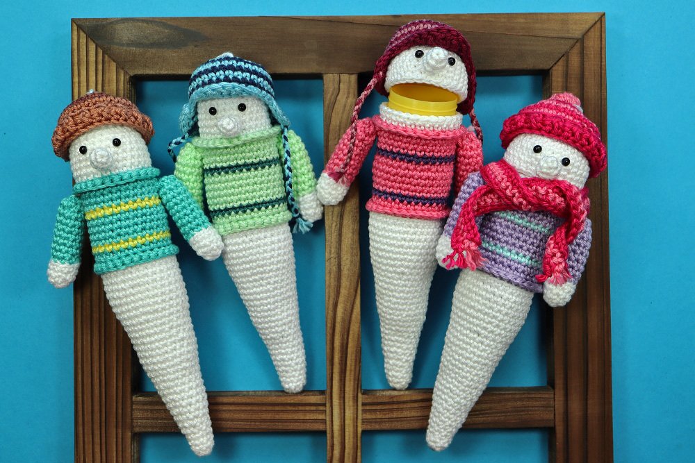 Reihe von vier gehäkelten Amigurumi-Flaschenhüllen mit winterlichen Pullovern und Mützen, nebeneinander hängend vor blauem Hintergrund.