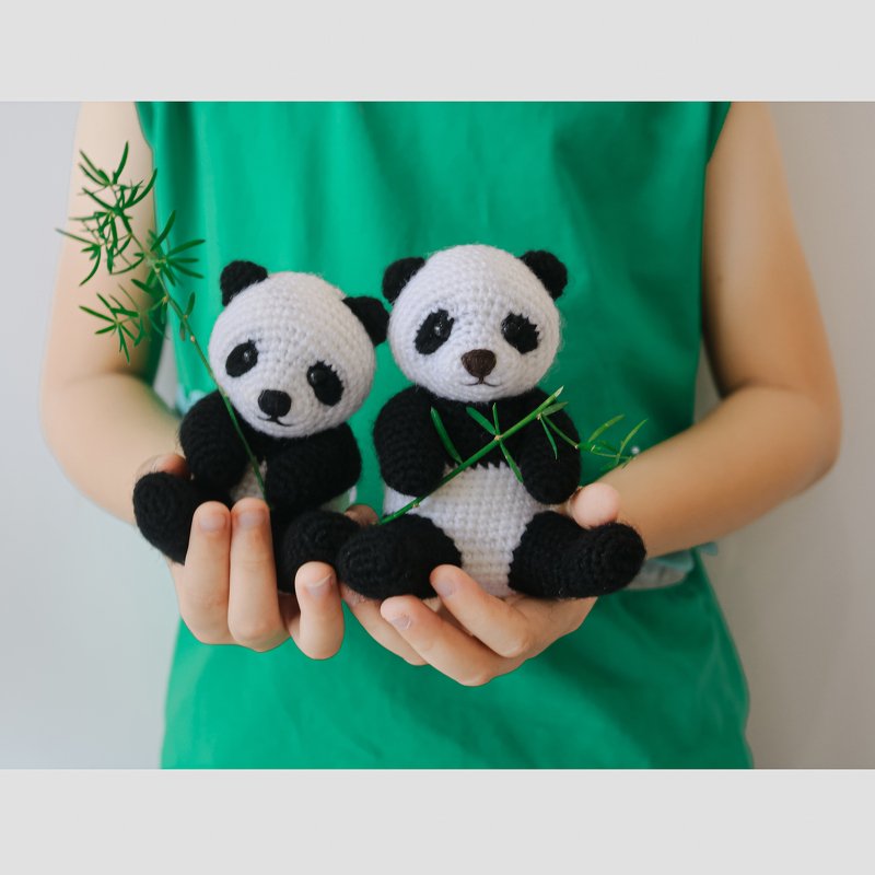 Pair of black-and-white crocheted panda amigurumi, each holding a small bamboo sprig, held in a person's hands.