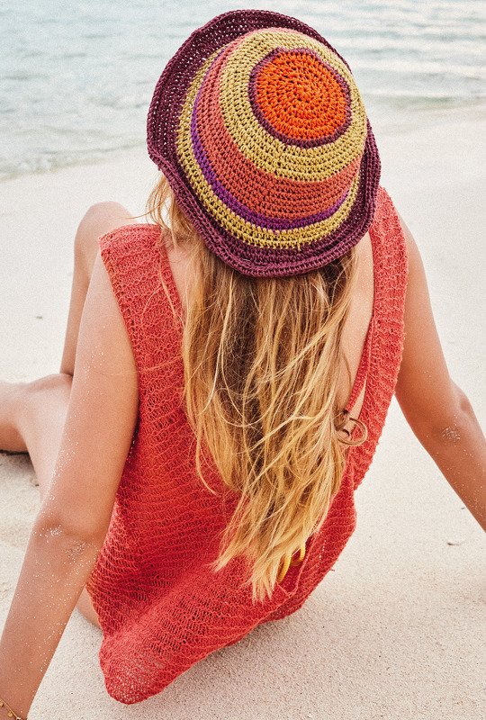 Rückenansicht einer Frau am Strand mit langem blondem Haar, trägt eine gehäkelte kreisförmige Mütze in Orange, Gelb, Lila und Burgunder