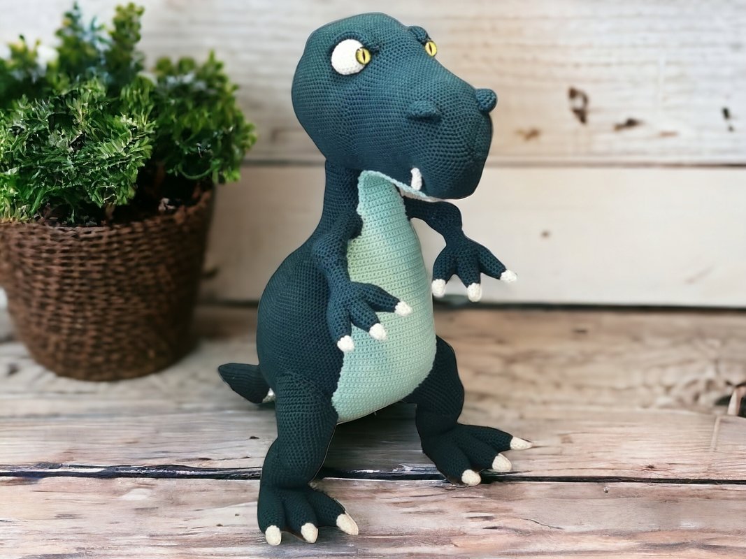 Gehäkelter Amigurumi
-T
-Rex in dunkelgrün mit mintfarbenem Bauch und weißen Krallen, stehend auf Holzbrett