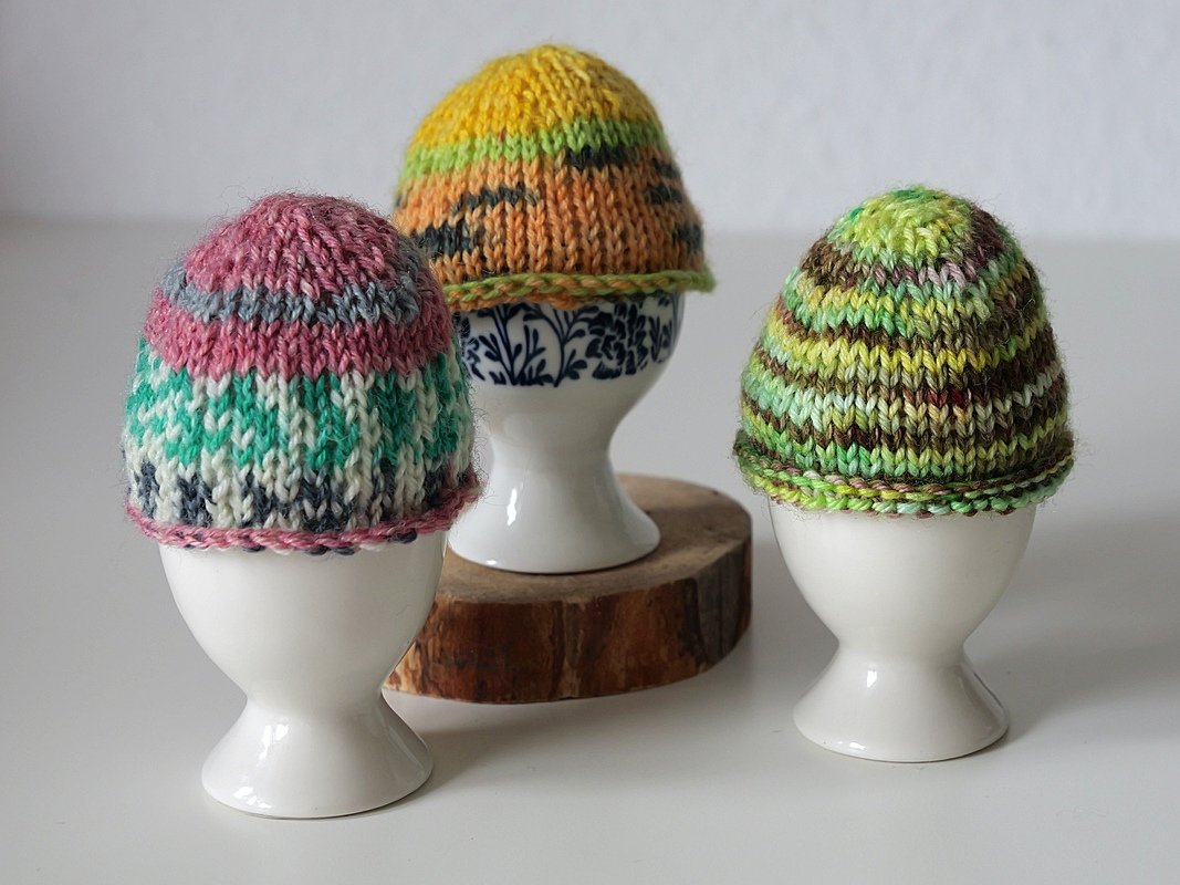 Drei gestrickte Eierwärmer mit unterschiedlichen Streifenmustern auf Eierbechern