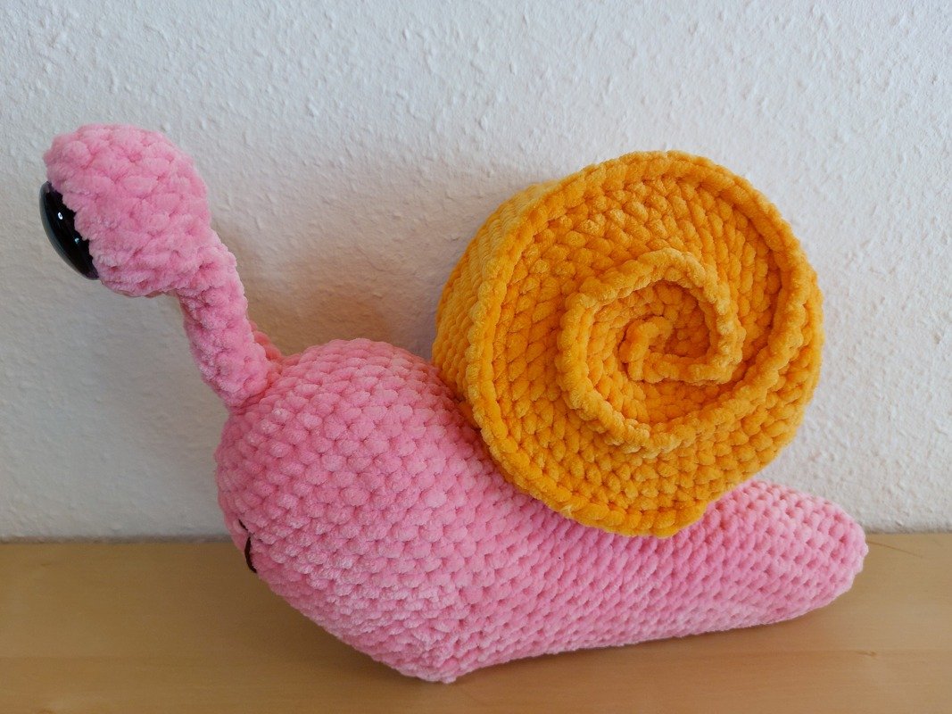 Gehäkelte Amigurumi‑Schnecke mit rosa Körper und orangefarbener Spirale auf hellem Holztisch vor weißer Wand