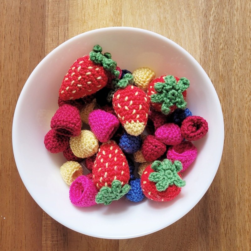 Weiße Schüssel mit gehäkelten roten Erdbeeren, pinken Himbeeren und kleinen blauen und gelben Häkelstücken auf Holzuntergrund