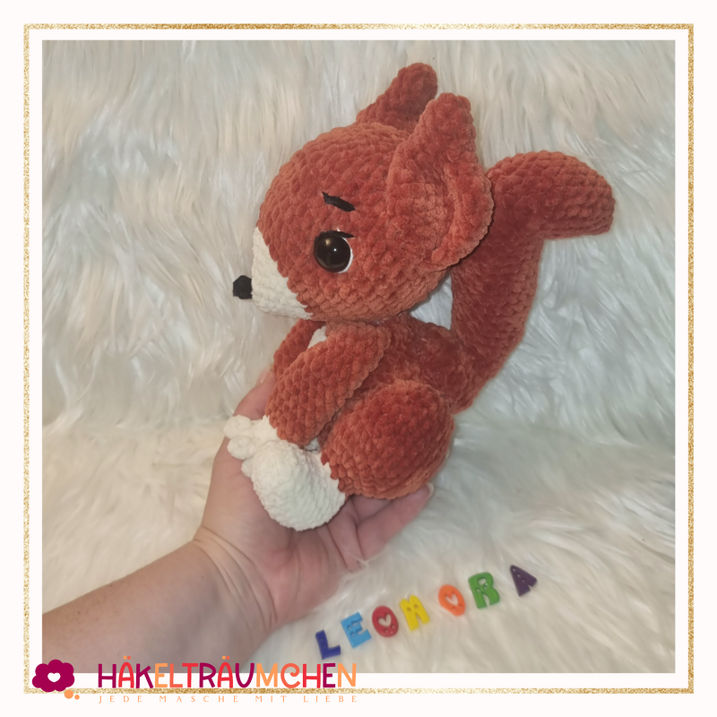 Gehäkeltes rotbraunes Eichhörnchen-Amigurumi mit weißer Schnauze und buschigem Schwanz, sitzend auf einer Hand