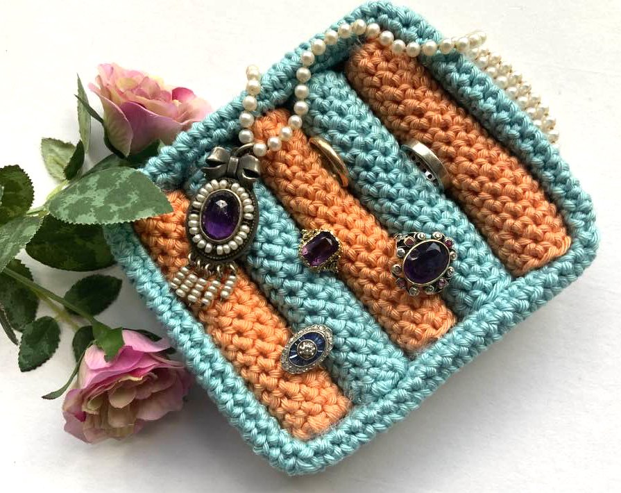 Gehäkelter Schmuck-Organizer in Türkis und Orange mit Ringen, Anhänger und Perlenkette, daneben Rosen.
