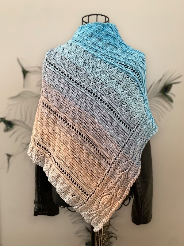 Blue-to-peach ombre crocheted triangle shawl with lace motifs, textured stitches, eyelet rows, and ruffled edge on a mannequin.