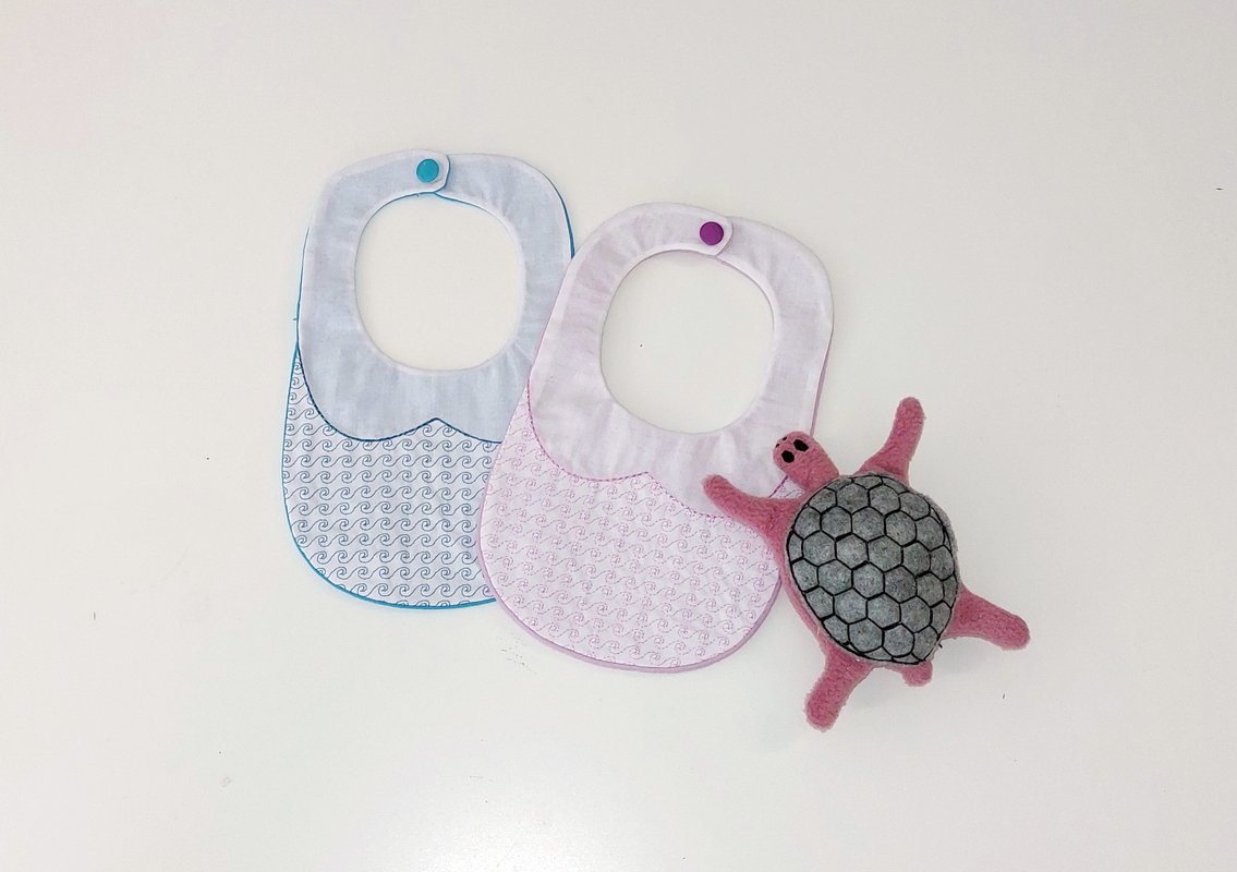 Zwei bestickte Baby-Lätzchen in Blau und Rosa mit Druckknopfverschluss neben einer gefilzten Schildkröte