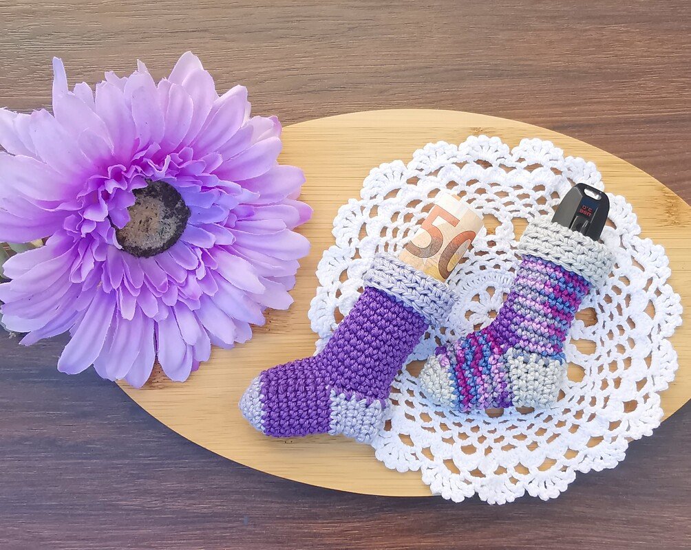 Two small purple crocheted sock pouches on a white doily on a wooden board; one contains a folded banknote and the other an electronic key fob, with a purple flower beside them.