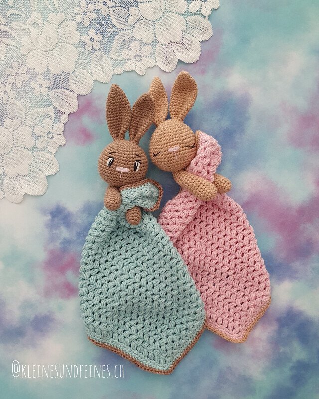 Two crocheted bunny loveys — mint green and pink blanket bodies with brown amigurumi bunny heads on a pastel background with lace.
