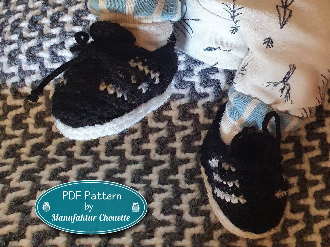 Gehäkelte Babysneaker schwarz mit weißer Sohle auf gemustertem Untergrund