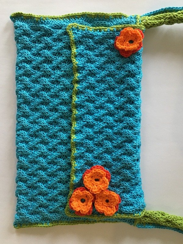 Türkisfarbene gestrickte Tasche mit Zopfmuster, limettengrüner Kante und vier orangefarbenen Häkelblüten.