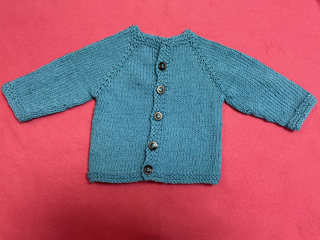 Türkise gestrickte Babyjacke mit Knopfleiste und Raglanärmeln auf pinkem Untergrund