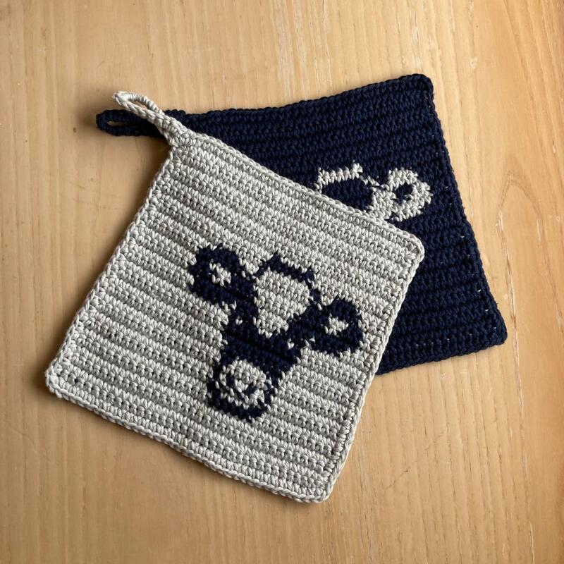 Zwei gehäkelte Topflappen in Hellgrau und Dunkelblau mit Kuhkopf-Motiv auf Holz.