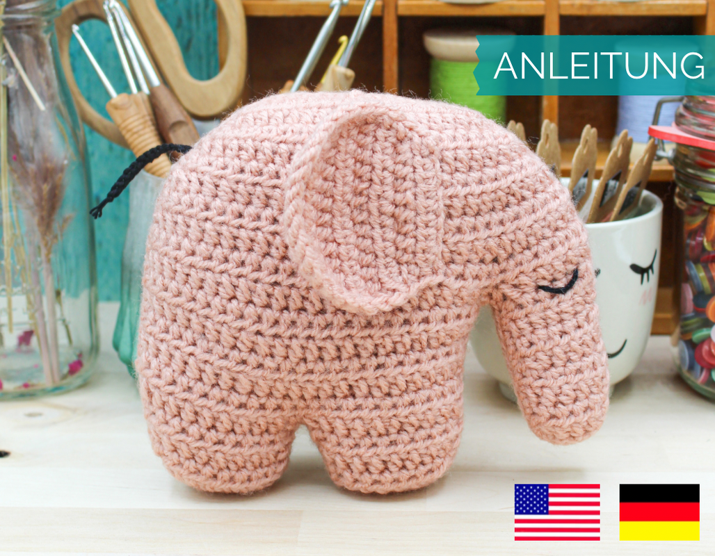 Altrosa gehäkelter Amigurumi‑Elefant mit geschlossenen Augen und großen Ohren vor Bastelutensilien; grüner ‚ANLEITUNG‘‑Banner oben rechts