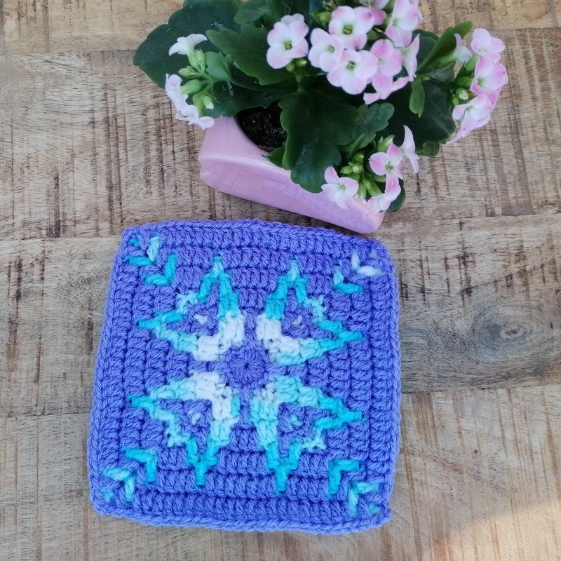 Gehäkeltes lila Quadrat mit weiß-türkisfarbenem Sternmotiv auf Holztisch neben Topfpflanze