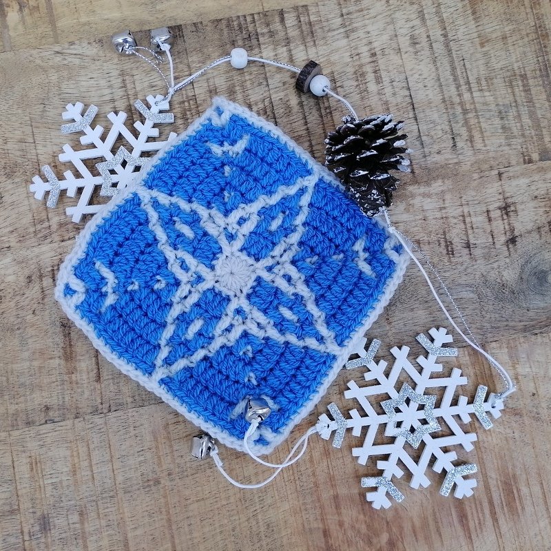 Gehäkeltes blaues Quadrat mit weißer Schneeflocke, Holzschneeflocken, Glöckchen und Zapfen auf Holzuntergrund