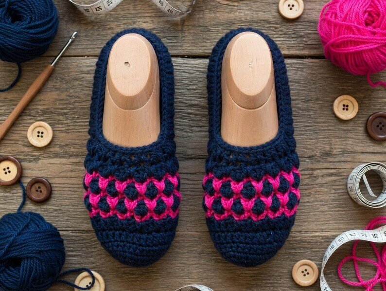 Top view of men's navy crochet slippers with pink band, surrounded by yarn balls, crochet hook, measuring tape and wooden buttons