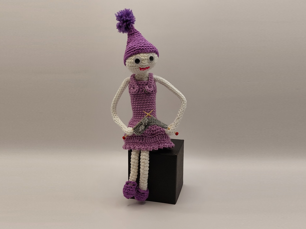 Gehäkelte Amigurumi-Puppe in Lila mit Bommelmütze, sitzend auf schwarzem Würfel, hält kleines Garnprojekt.