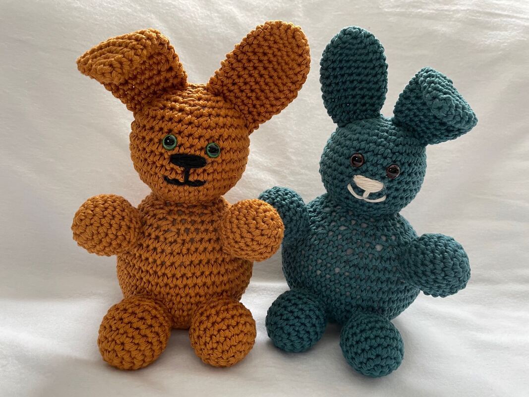 Zwei gehäkelte Amigurumi-Hasen in Orange und Petrol sitzen nebeneinander vor weißem Hintergrund.