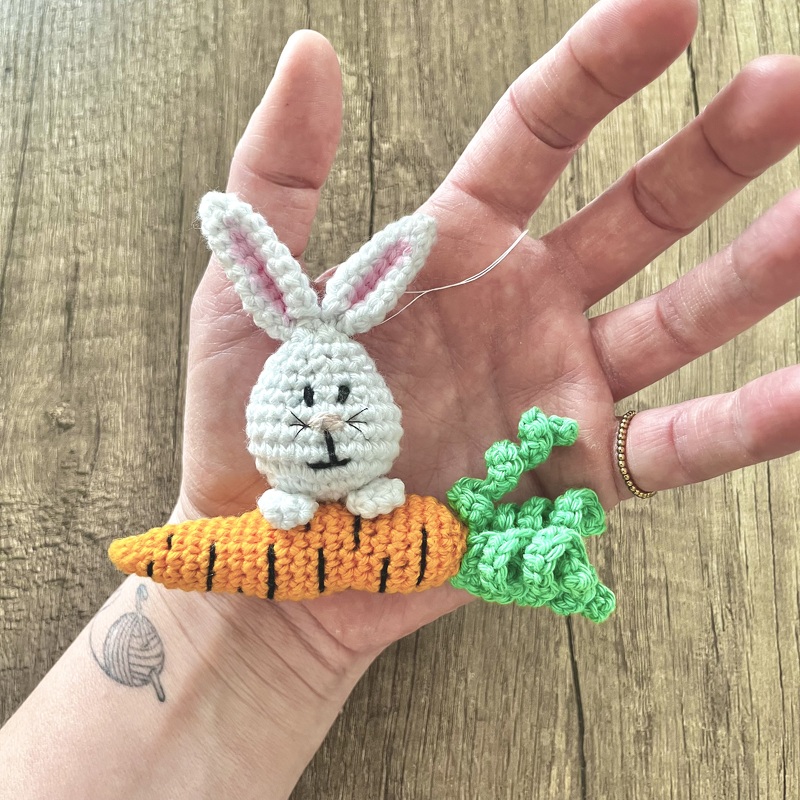 Weißer gehäkelter Amigurumi-Hase auf orangefarbener Möhre mit grünen Blättern, in einer Hand gehalten