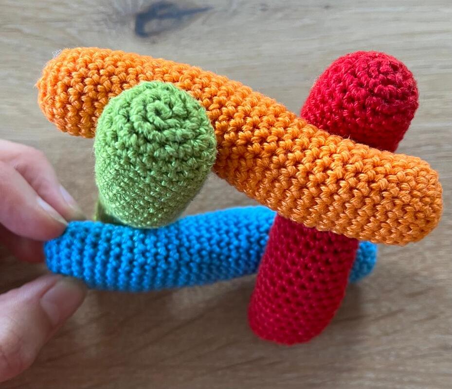Vier gehäkelte Röhren in Rot, Orange, Blau und Grün; eine Hand hält das blaue Element
