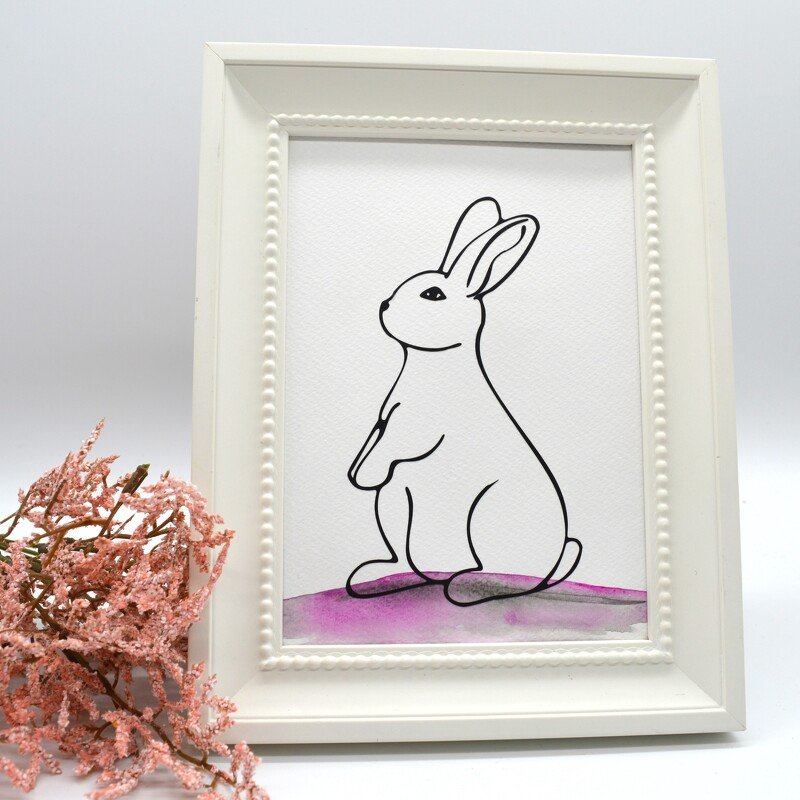 Einfaches Linienmotiv eines sitzenden Hasen in weißem Rahmen auf hellem violetten Aquarellfleck, links getrocknete rosa Zweige