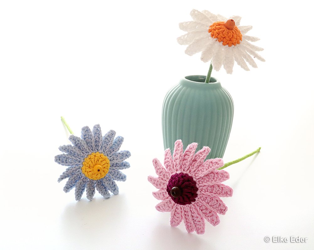 Drei gehäkelte Gänseblümchen (weiß, hellblau, rosa) auf weißem Hintergrund; eines steht in einer mintgrünen Vase.