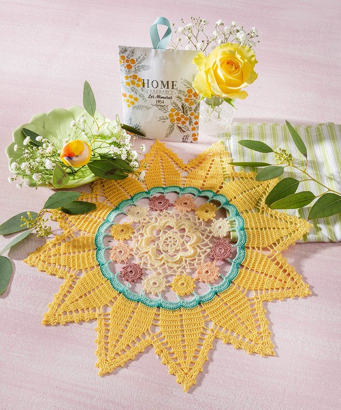 Sternförmiges gelbes Häkeldeckchen mit pastellfarbener Blumenmitte auf rosa Untergrund, daneben gelbe Rose und grüne Zweige
