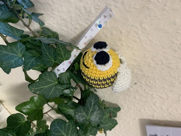 Gehäkelte gelb‑graue Biene mit weißen Flügeln hängt am Efeu vor heller Wand.