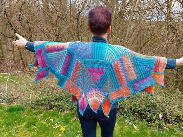 Rückansicht einer Person, die ein farbenfrohes Stricktuch mit geometrischen Dreiecken und ausgezacktem Saum im Freien trägt.