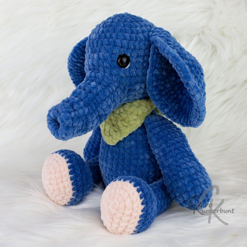 Gehäkelter blauer Amigurumi-Elefant mit rosa Fußsohlen und grünem Halstuch auf hellem Hintergrund