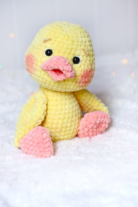 Plush yellow crochet duckling with pink beak, pink feet and blushed cheeks seated on white fabric