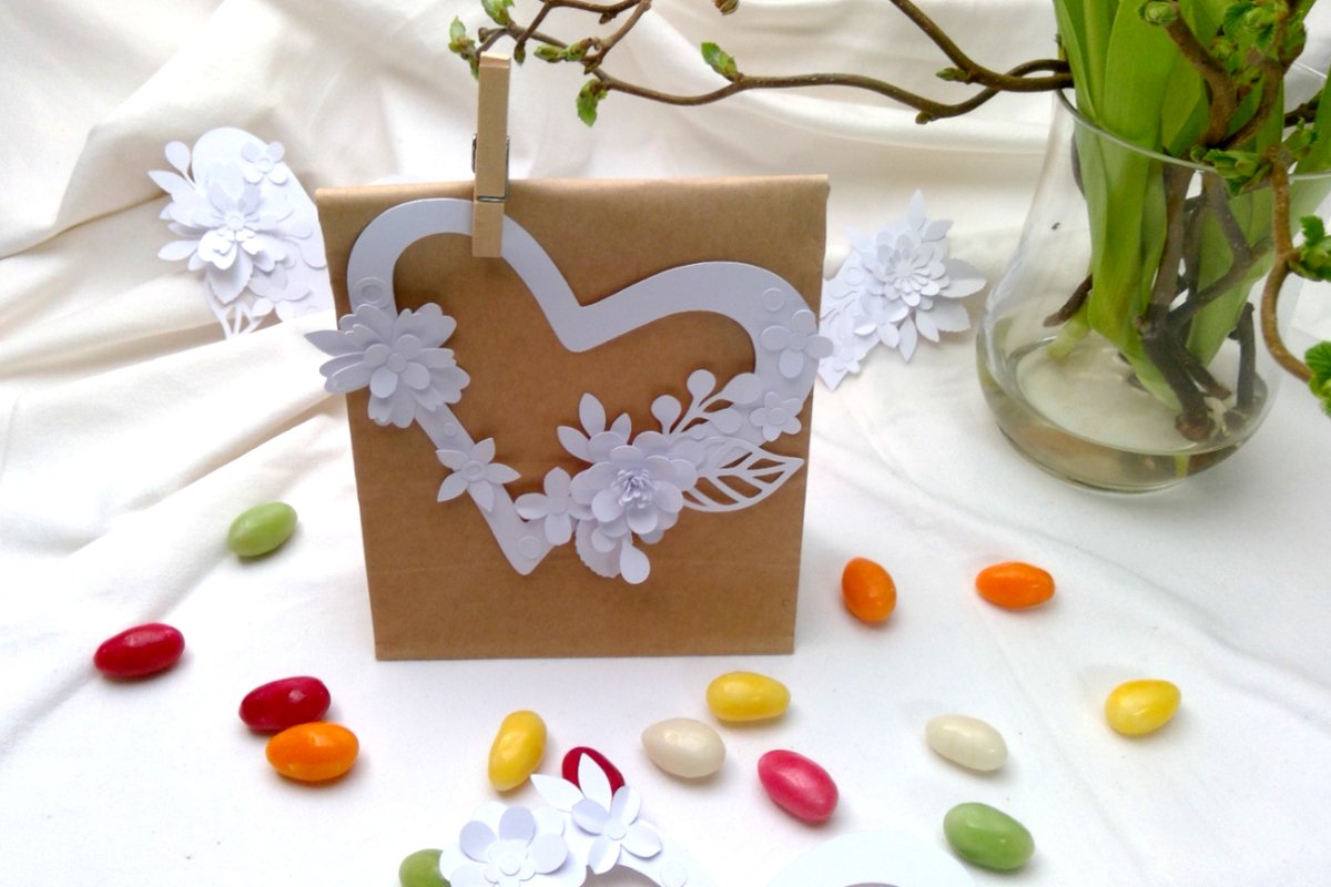 Braune Geschenktüte mit weißem, filigran geschnittenem Papierherz mit Blumen, umgeben von bunten Süßigkeiten.