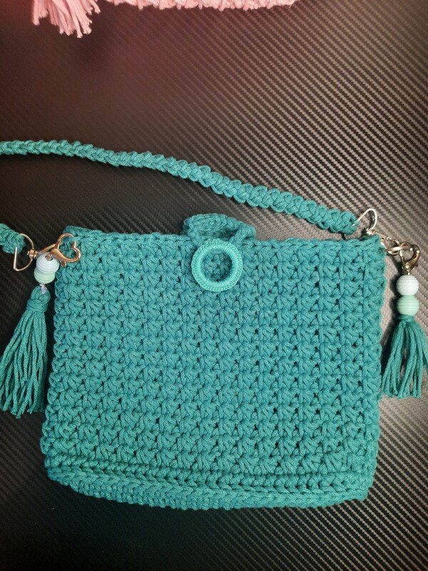 Türkise gehäkelte Umhängetasche mit geflochtenem Riemen, Quasten, Perlen und ringförmigem Verschluss auf dunklem Hintergrund.