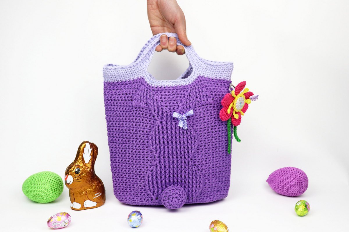 Lila gehäkelte Tasche mit Hasenmuster und Bommel, gehalten von einer Hand, umgeben von Ostereiern und Schokoosterhase