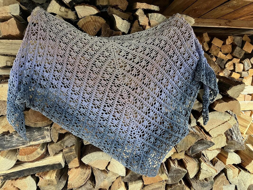 Gehäkeltes Dreieckstuch in Farbtönen Rosé, Blaugrau und Dunkelblau mit offenem Lochmuster, auf Holzstapel drapiert.