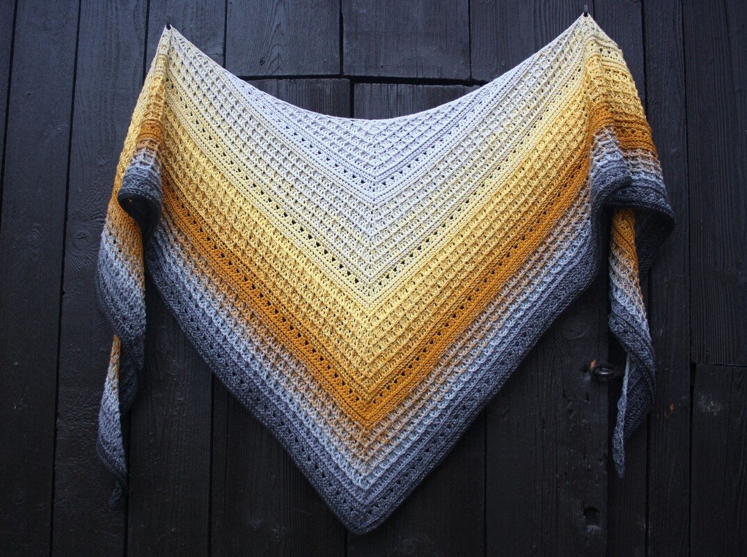 Gehäkeltes Dreieckstuch mit Verlaufsgarn in Creme, Gelb und Grau vor dunkler Holzwand